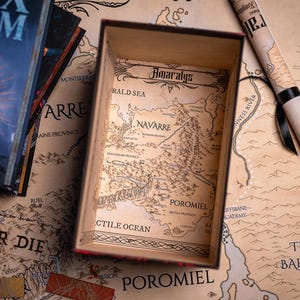 May include: A wooden box with a map of Amaralys inside, next to a rolled-up map and books. The box and maps have a vintage aesthetic with detailed illustrations. The books have the title "Storm" on the cover.