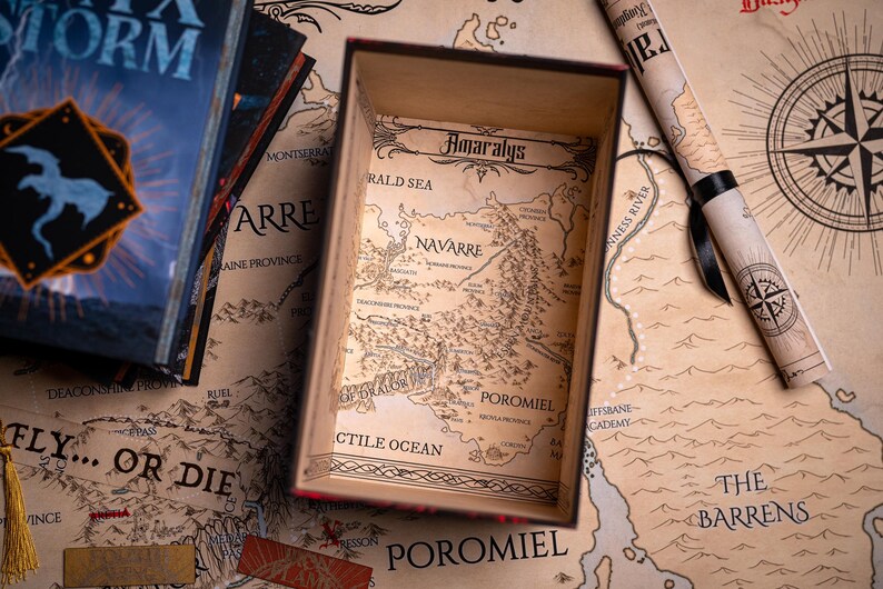 May include: A wooden box with a map of Amaralys inside, next to a rolled-up map and books. The box and maps have a vintage aesthetic with detailed illustrations. The books have the title "Storm" on the cover.