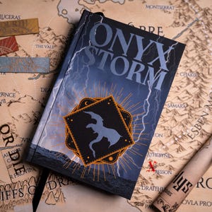 May include: A hardcover book titled "ONYX STORM" with a lightning bolt design. The book has a black and orange emblem with a silhouette. The image includes bookmarks, a map, and a scroll.
