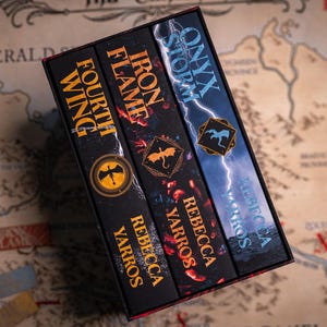May include: A boxed set of three fantasy novels by Rebecca Yarros. The book spines are black with gold and red lettering. Titles include "Fourth Wing", "Iron Flame", and "Onyx Storm". Each book features a unique emblem. The set rests on a map.
