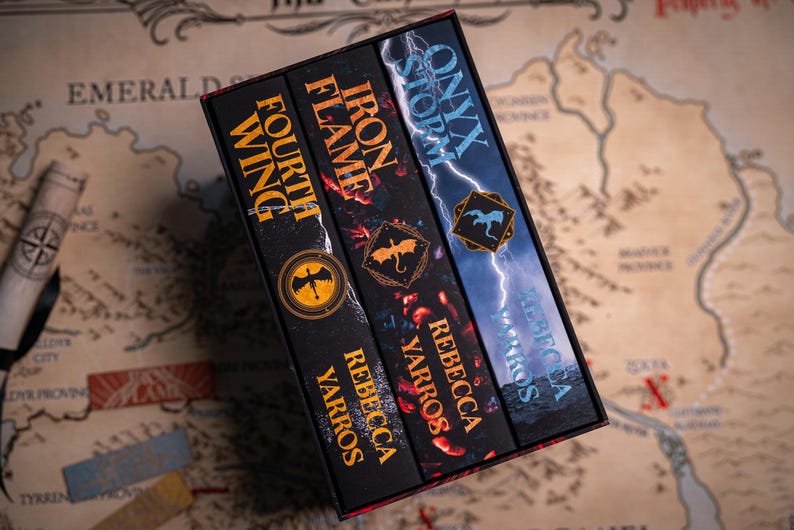 May include: A boxed set of three fantasy novels by Rebecca Yarros. The book spines are black with gold and red lettering. Titles include "Fourth Wing", "Iron Flame", and "Onyx Storm". Each book features a unique emblem. The set rests on a map.