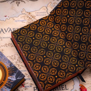 May include: An open, black journal with a repeating gold geometric pattern and a brown spine rests on a map. A book with a dragon emblem is visible on the left. The journal's pages are visible.