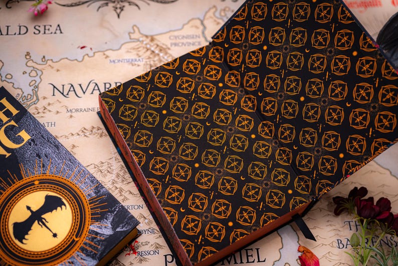 May include: An open, black journal with a repeating gold geometric pattern and a brown spine rests on a map. A book with a dragon emblem is visible on the left. The journal's pages are visible.