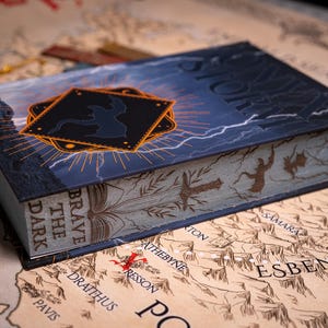 May include: A hardcover book titled "STORM" with a dark blue cover featuring lightning and a black emblem. The book's spine reads "BRAVE THE DARK". The book is resting on a map with various place names.