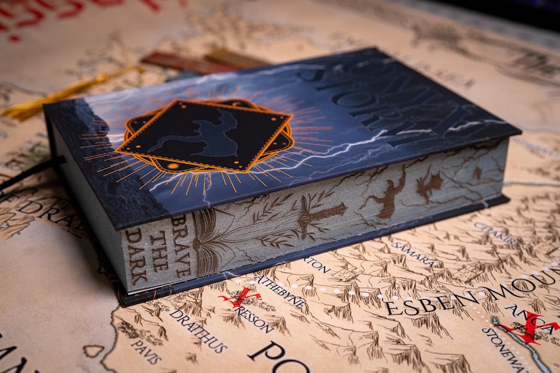 May include: A hardcover book titled "STORM" with a dark blue cover featuring lightning and a black emblem. The book's spine reads "BRAVE THE DARK". The book is resting on a map with various place names.