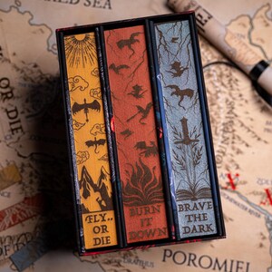 May include: A boxed set of three books with decorative covers. The covers are yellow, red, and blue, each with a unique design featuring dragons and text. The box sits on a map with the words "FLY... OR DIE". A rolled-up map is in the background.