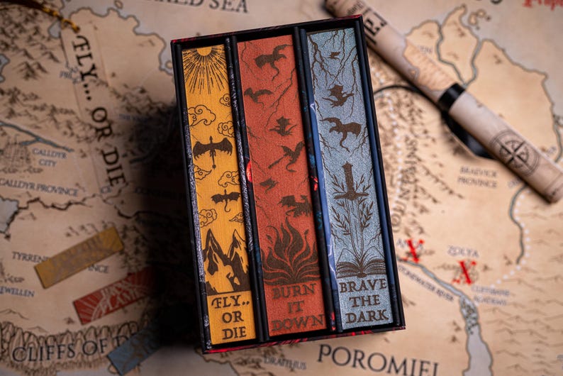 May include: A boxed set of three books with decorative covers. The covers are yellow, red, and blue, each with a unique design featuring dragons and text. The box sits on a map with the words "FLY... OR DIE". A rolled-up map is in the background.