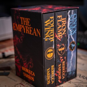 May include: A boxed set of fantasy novels titled "The Empyrean" by Rebecca Yarros. The set includes "Fourth Wing", "Iron Flame", and "Onyx Storm". The books have black covers with orange and red accents. The box and books are on a map.