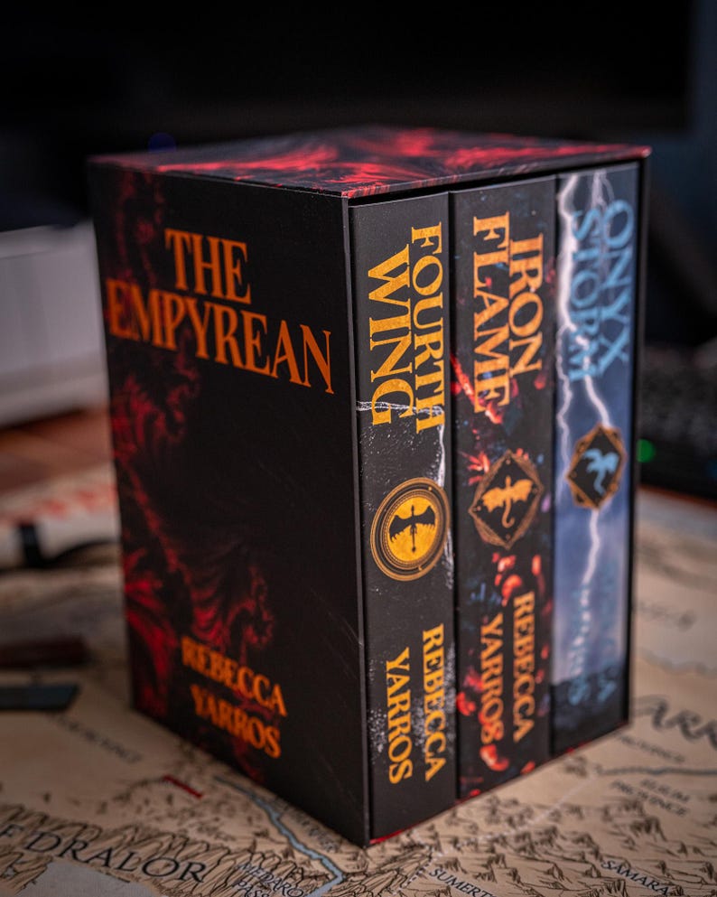 May include: A boxed set of fantasy novels titled "The Empyrean" by Rebecca Yarros. The set includes "Fourth Wing", "Iron Flame", and "Onyx Storm". The books have black covers with orange and red accents. The box and books are on a map.