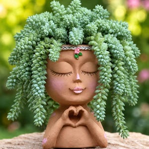 May include: A terracotta-colored planter shaped like a human head with closed eyes, pink cheeks, and hands forming a heart shape at the chest. A silver braided headband with a small pink flower adorns the forehead, topped with a trailing succulent.