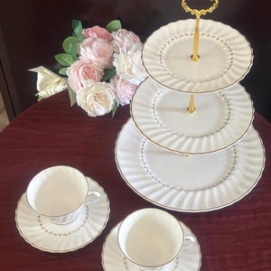 May include: A three-tiered serving stand with gold accents and matching teacups and saucers. The white porcelain pieces have a gold trim and a delicate leaf pattern. A bouquet of pink and white flowers sits nearby.