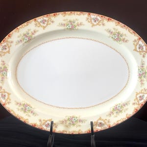 May include: An oval serving platter with a cream-colored background and a gold and red floral border. The platter is decorated with a delicate floral pattern in shades of pink, yellow, and green.