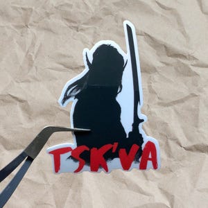 May include: Black and white silhouette of a person holding a sword with the word "TSK'VA" in red letters.