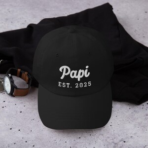 May include: Black baseball cap with the word "Papi" embroidered in white script, with "EST. 2025" below. A watch with a brown leather band is visible to the left of the cap.