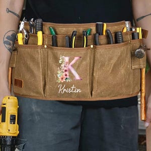 May include: A brown canvas tool belt with multiple pockets, filled with various tools. The belt features a floral design with the letter "K" and the name "Kristin" printed on the front. A yellow drill is visible on the left.