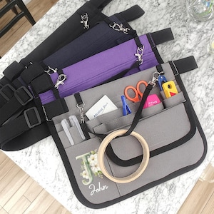 May include: A grey craft apron with multiple pockets, holding scissors, pens, and other tools. The apron has a black strap and trim, with a wooden embroidery hoop attached. The apron is personalised with the name "John" and the initial "J".