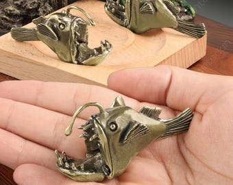 May include: A pair of gold-colored anglerfish figurines. The figurines are detailed and realistic, with sharp teeth and a lure on top of their heads. One figurine is held in a hand, while the other is on a wooden surface.