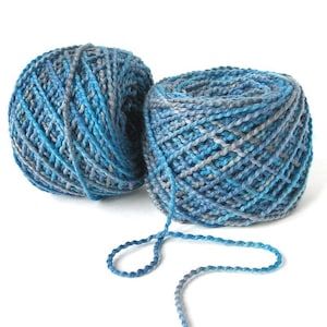 May include: Two balls of yarn in shades of blue and gray sit on a white background. The yarn is tightly wound, with visible texture and a slight sheen. One ball has a strand of yarn extending from it, suggesting it's ready for use in a knitting or crochet project.