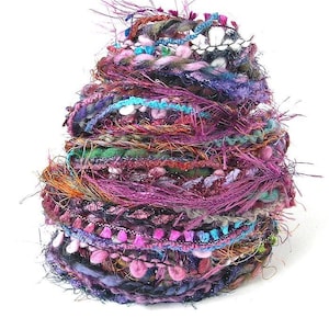 May include: A colorful, textured yarn ball with a variety of threads and embellishments. The yarn features shades of purple, pink, green, and blue, with a mix of textures including fringe, beads, and metallic threads. This craft supply is ideal for knitting and crochet projects.