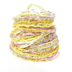 May include: A vibrant, layered yarn cake in shades of yellow, pink, and white. The yarn has a textured appearance with various threads and ribbons, creating a visually appealing and tactile product. Ideal for crafting projects.