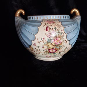 May include: A ceramic bowl featuring a cream-coloured centre with a multi-coloured floral pattern, flanked by two light blue, fan-shaped side panels with gold-toned handles. The top rim has a perforated design and gold-toned trim accents.