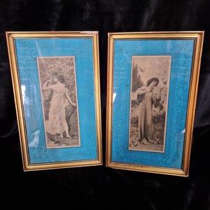 May include: Two framed vintage prints. Each print features a woman in a flowing gown. The prints are set against a textured, turquoise background within gold-colored frames. The frames are rectangular and have a beveled edge.