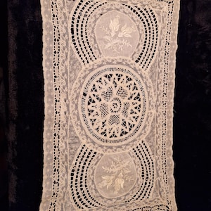 Peut inclure: Nappe en dentelle rectangulaire blanc cassé avec un motif floral délicat. La nappe présente trois médaillons circulaires avec broderie florale et motifs ajourés complexes. Les bords sont festonnés, ajoutant à son charme vintage.