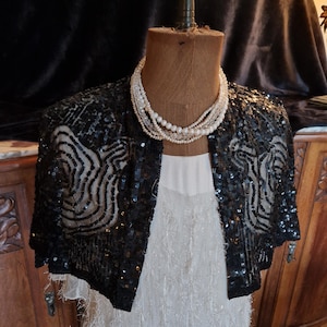 May include: A black sequined bolero jacket with a zebra-like pattern is displayed on a mannequin. A multi-strand pearl necklace and a white dress with fringe detail complete the ensemble. The background features a dark velvet drape and antique furniture.