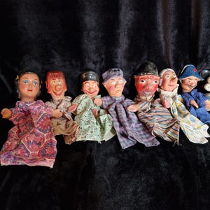 May include: A group of seven vintage hand puppets with painted faces and various outfits. The puppets include a Mickey Mouse-like character and others in dresses, hats, and striped shirts. They are displayed on a dark, textured background.
