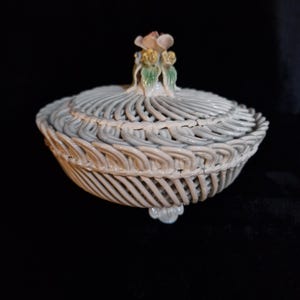 May include: A white ceramic basket with a lid, featuring a woven design. The lid is topped with a floral accent in shades of pink, green, and yellow. The basket has a round shape and sits on three small feet.