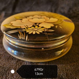 Puede incluir: Una caja de joyería redonda, negra y dorada con un diseño floral. La tapa presenta una flor blanca con detalles dorados sobre un fondo negro. La caja tiene un diámetro de 13 cm y pesa 679 gramos.