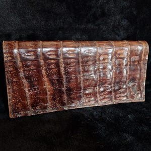 Può includere: Un portafoglio in pelle marrone con una texture a pelle di coccodrillo. Il portafoglio è rettangolare e ha una finitura lucida. Il colore è un marrone scuro intenso con riflessi marroni più chiari. Il portafoglio è chiuso e sembra vuoto.