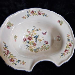 Può includere: Piatto decorativo in ceramica bianca con bordo smerlato e bordo blu. Il piatto presenta un motivo floreale e di uccelli in rosa, giallo, blu e verde. È raffigurata anche una farfalla. Il piatto ha una forma unica con un'indentazione curva.
