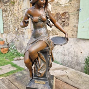 May include: Bronze sculpture of a seated woman holding a circular object. The sculpture is on a pedestal with the word "JEUNESSE" inscribed. The woman is draped in flowing fabric, with her hair cascading down her shoulder. The sculpture is placed on a wooden surface.