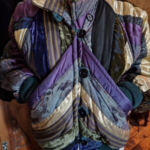 Puede incluir: Chaqueta acolchada con diseño patchwork. Presenta una mezcla de texturas y colores, incluyendo dorado, morado, verde y azul. La chaqueta tiene grandes botones negros y puños de canalé. Una prenda de abrigo única.