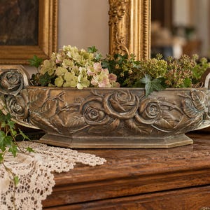 May include: An elongated, bronze-toned planter featuring a detailed relief of sculpted roses and leaves along its exterior. The vessel has curved, raised ends and a flat base, currently holding a cluster of green ivy and light-coloured flowers.