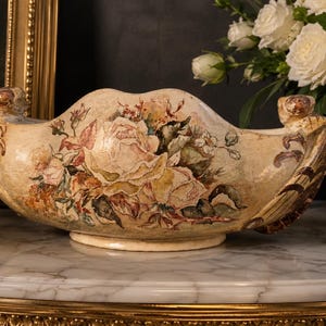 May include: An oval-shaped ceramic bowl with a cream-coloured base, featuring a painted floral design of large roses in shades of pink, yellow, and green. The bowl has a scalloped rim and decorative handles on each side with brown accents.