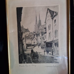 Peut inclure: Une gravure encadrée en noir et blanc représentant une scène de rue à Chartres, intitulée Chartres Rue du Bourg. L'œuvre montre une rue pavée avec des charrettes tirées par des chevaux, des bâtiments historiques et une cathédrale à deux flèches.