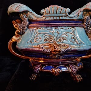 Peut inclure: Un récipient en céramique antique orné, de couleur bleu clair, violet et or. Le récipient présente des sculptures en relief détaillées, des poignées et des pieds décoratifs. Le design comprend un visage et des motifs floraux.