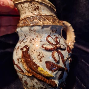 Peut inclure: Pichet en céramique avec une base bleu clair et des accents marron. Le pichet présente un motif d'oiseau et de fleurs. La poignée a un motif texturé. Le bord supérieur est de couleur marron foncé.