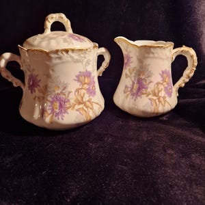 Peut inclure: Un sucrier vintage en porcelaine avec couvercle et une crémière. Les deux pièces sont blanches avec des motifs floraux violets et dorés. Le sucrier a une poignée sur le couvercle et deux poignées latérales. La crémière a une anse incurvée et un bec verseur.