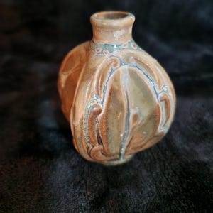 Peut inclure: Petit vase artisanal en céramique, corps bulbeux et col étroit. Il présente un motif de couleurs marron et beige avec des accents bleus et des motifs floraux décoratifs en relief. Le vase est probablement à des fins décoratives.