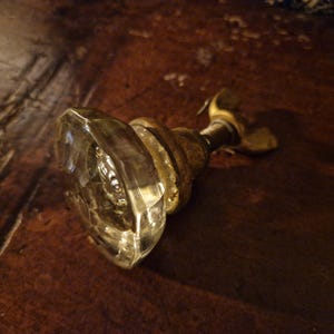 Op de afbeelding: Antieke glazen deurknop met een facetgeslepen, heldere glazen kop en een messingkleurige metalen basis. De deurknop ligt op een donker houten oppervlak en toont zijn vintage design en vakmanschap. Een klassiek stuk voor de interieurdecoratie.