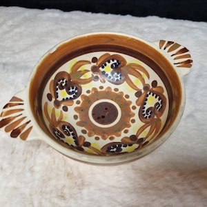 Puede incluir: Un tazón de cerámica blanca con dos asas. El tazón está decorado con un patrón floral marrón, amarillo y azul.