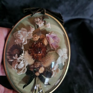 May include: An oval-shaped, vintage floral display encased in glass. The arrangement features dried flowers in shades of cream, brown, and pink, tied with a black ribbon. The display is framed in gold and has a dark green ribbon for hanging.