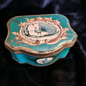 Peut inclure: Une boîte à bijoux vintage de couleur turquoise avec une illustration de paon sur le couvercle. La boîte a une bordure dorée et un design décoratif autour de l'image du paon. La boîte est probablement en métal.