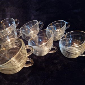 Peut inclure: Ensemble de huit tasses à thé en verre transparent avec des anses. Les tasses présentent un motif texturé et tourbillonnant autour du bol. Elles sont disposées sur une surface sombre, contrastant avec le verre transparent.