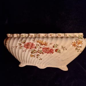 Könnte beinhalten: Cremefarbener, länglicher Pflanztopf mit floralem Design. Der Pflanztopf hat einen geriffelten Körper, einen gewellten Rand und drei kleine Füße. Das florale Muster umfasst rote, gelbe und grüne Blumen und Blätter.