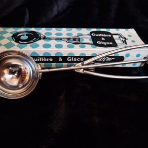 Peut inclure: Une cuillère à glace vintage avec une cuvette et une poignée argentées brillantes. La cuillère est à côté d'une boîte à pois turquoise et blancs avec l'inscription "Cuillère à Glace".