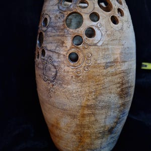 Peut inclure: Un vase en céramique beige fait à la main avec un design unique. Le vase présente une série de découpes circulaires de différentes tailles sur la partie supérieure. La surface a un aspect texturé et vieilli avec des teintes marron et bleu.